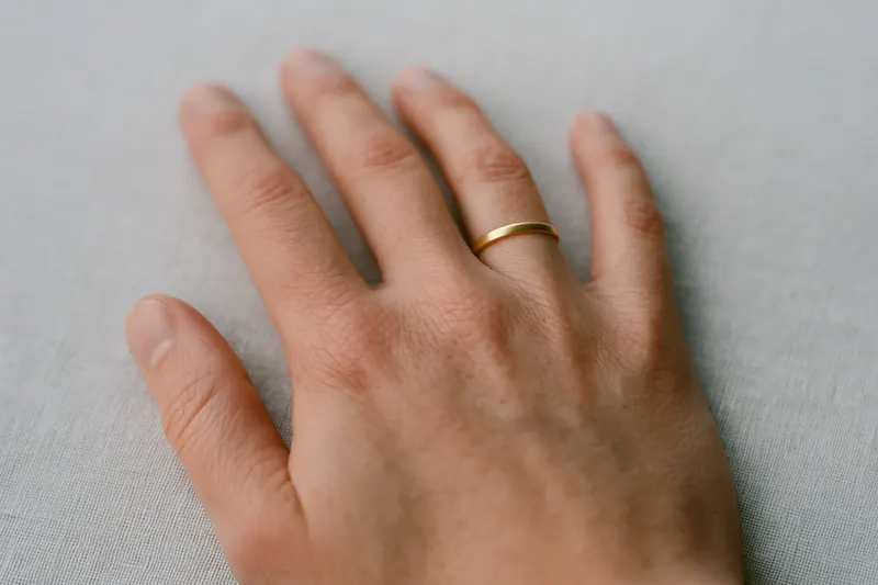A hand held palm down on pale linen with a plain gold band pulled partway off the ring finger and stopped at the middle knuckle.