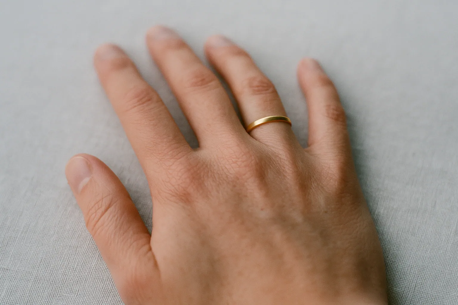 A hand held palm down on pale linen with a plain gold band pulled partway off the ring finger and stopped at the middle knuckle.