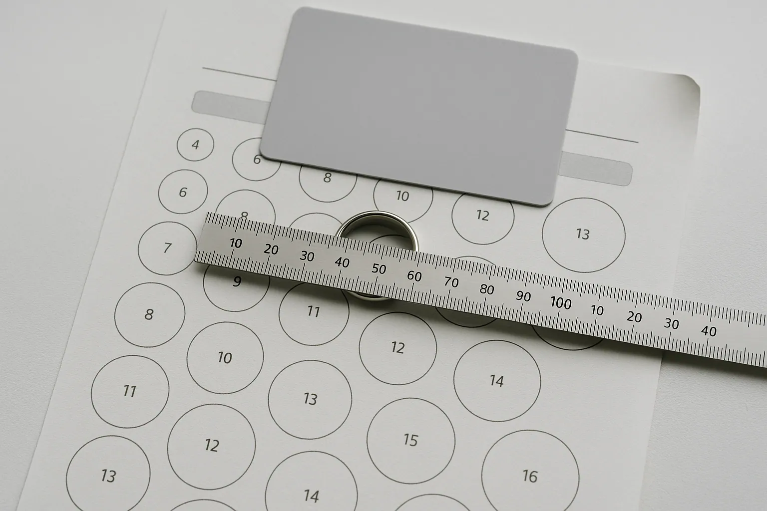 A steel millimeter ruler laid across the inside of a plain platinum band on a white desk, with a bank card and a printed page of numbered ring size circles underneath.